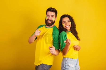 Photo for Couple of brazil soccer supporters, dressed in the colors of the nation, black woman, caucasian man. Pointing at camera. - Royalty Free Image