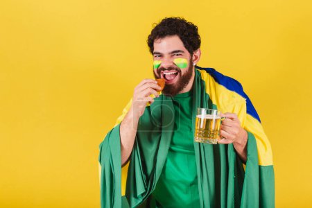 Téléchargez les photos : Homme caucasien avec barbe, brésilien, fan de football brésilien, tenant un verre de bière et un pilon de poulet, nourriture typique brésilienne. - en image libre de droit