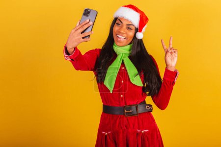Téléchargez les photos : Belle femme brésilienne noire, vêtue de vêtements de Noël, Père Noël claus, smartphone prenant autoportrait, selfie. - en image libre de droit