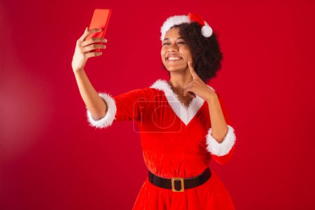 Téléchargez les photos : Belle femme brésilienne noire, habillée comme Santa Claus, maman Claus, en appel vidéo à l'aide d'un smartphone - en image libre de droit
