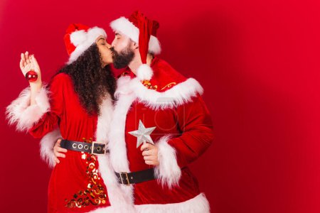 Photo for Brazilian couple, dressed for christmas, santa claus, mummy claus. holding christmas decoration, exchanging kiss. approved, black woman and caucasian man. - Royalty Free Image
