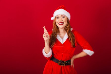 Photo for Beautiful Brazilian red-haired woman, dressed in Christmas clothes, Santa Claus. fingers crossed, wishing and hoping. - Royalty Free Image
