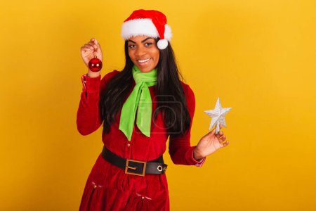 Photo for Beautiful black Brazilian woman, dressed in christmas clothes, santa claus, holding christmas decoration. star and ball. - Royalty Free Image