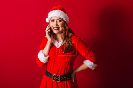 Photo for Beautiful Brazilian red-haired woman, dressed in Christmas clothes, Santa Claus. talking on voice call by smartphone. - Royalty Free Image