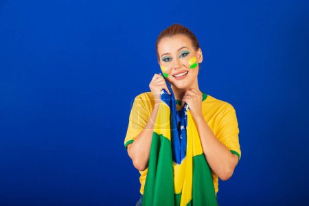 Téléchargez les photos : Femme caucasienne, rousse, fan de football brésilien, brésilien, fond bleu, souriante et heureuse avec le drapeau dans les mains - en image libre de droit