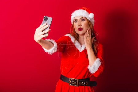 Téléchargez les photos : Belle femme rousse brésilienne, vêtue de vêtements de Noël, le Père Noël. appel vidéo via smartphone. - en image libre de droit