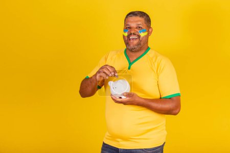 Foto de Hombre negro brasileño, fanático del fútbol de Brasil. tener cerdito y moneda, economía, jubilación. ahorros. - Imagen libre de derechos