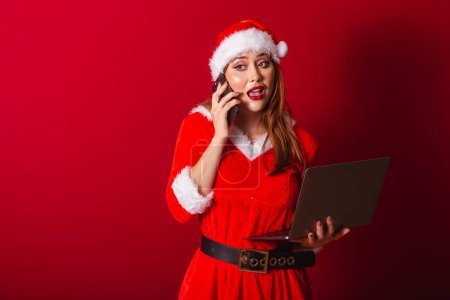 Téléchargez les photos : Belle femme rousse brésilienne, vêtue de vêtements de Noël, le Père Noël. tenant un ordinateur portable et un smartphone, occupé. - en image libre de droit