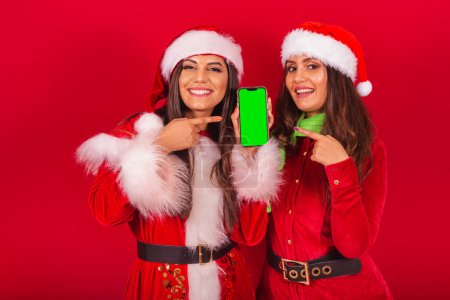 Téléchargez les photos : Des amies brésiliennes vêtues de Noël, le Père Noël. tenant téléphone portable avec écran chromatique vert. - en image libre de droit