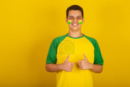 Foto de Joven aficionado al fútbol brasileño. vestido de verde, y amarillo, pulgares hacia arriba, como - Imagen libre de derechos