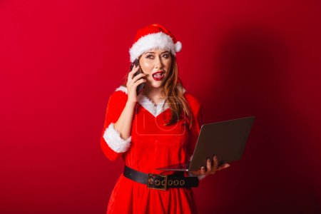 Photo for Beautiful Brazilian red-haired woman, dressed in Christmas clothes, Santa Claus. holding notebook and smartphone, busy. - Royalty Free Image