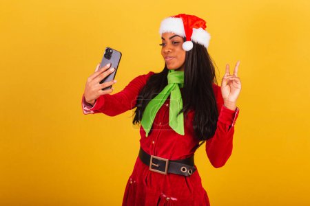 Foto de Hermosa mujer brasileña negra, vestida con ropa de Navidad, santa claus, teléfono inteligente tomando autorretrato, selfie. - Imagen libre de derechos