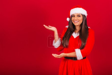 Photo for Beautiful Brazilian woman dressed in Christmas clothes, Santa Claus. showing something to the left. - Royalty Free Image