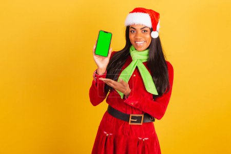 Téléchargez les photos : Belle femme brésilienne noire, vêtue de vêtements de Noël, Père Noël claus, smartphone et montrant écran chromé vert. - en image libre de droit