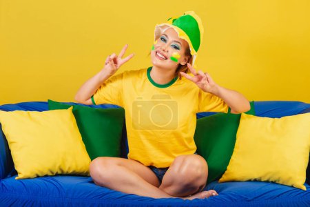 Photo for Caucasian woman, redhead, brazil soccer fan, brazilian, on the couch making peace and love symbol - Royalty Free Image