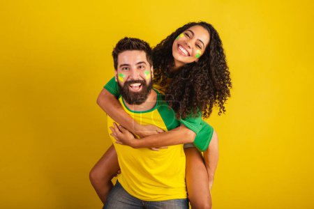 Foto de Pareja de aficionados al fútbol brasileño, vestidos con los colores de la nación, mujer negra, hombre caucásico. besando, amor. - Imagen libre de derechos