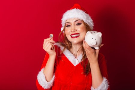 Photo for Beautiful Brazilian red-haired woman, dressed in Christmas clothes, Santa Claus. holding piggy bank and coin. - Royalty Free Image
