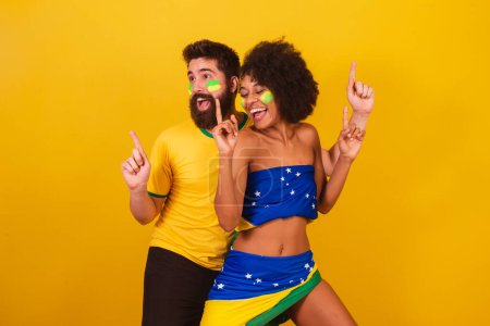 Téléchargez les photos : Couple de fans de football brésilien, habillé dans les couleurs du brésil, femme noire, homme caucasien. danser ensemble, s'amuser. - en image libre de droit