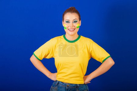 Foto de Mujer caucásica, pelirroja, aficionada al fútbol brasileño, brasileña, fondo azul, manos en la cintura - Imagen libre de derechos