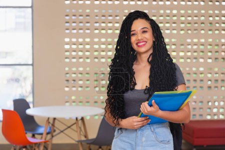 Téléchargez les photos : Jeune femme noire, brésilienne, étudiante dans le couloir du collège, se préparant à aller en cours, souriante. - en image libre de droit