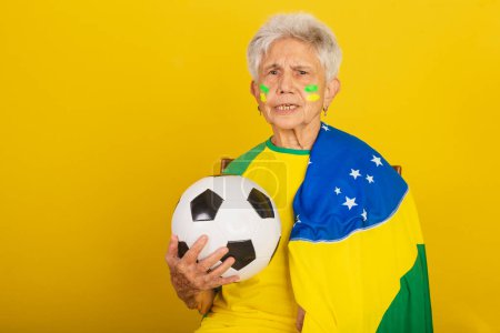 Téléchargez les photos : Femme âgée, fan de football du Brésil, acclamant avec le drapeau. tenant le ballon de football - en image libre de droit