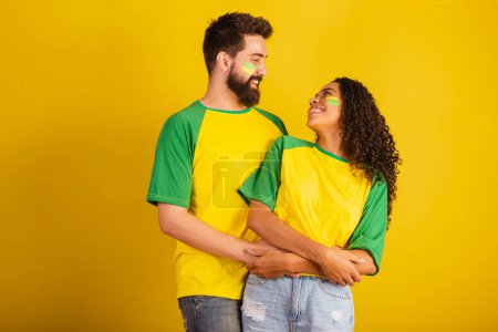 Téléchargez les photos : Couple de supporters brésiliens de football, vêtus aux couleurs de la nation, femme noire, homme caucasien. embrassé. - en image libre de droit