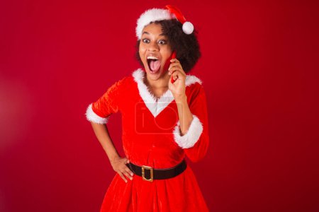 Photo for Beautiful black brazilian woman, dressed as santa claus, mama claus, talking on voice call by smartphone, surprised, wow, amazing - Royalty Free Image