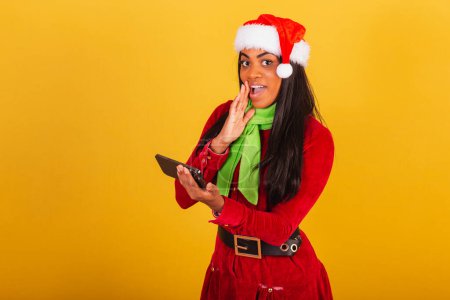 Photo for Beautiful black Brazilian woman, dressed in Christmas clothes, Santa Claus, smartphone and celebrating. - Royalty Free Image