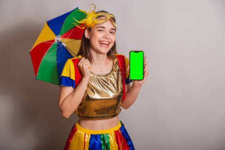 Photo for Beautiful Brazilian, Caucasian woman, wearing frevo, carnival clothes, wearing a mask. holding smartphone with green screen in chroma. - Royalty Free Image