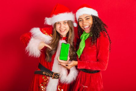 Téléchargez les photos : Des amies brésiliennes, avec des vêtements de Noël, le Père Noël. écran montrant smartphone avec chroma. - en image libre de droit