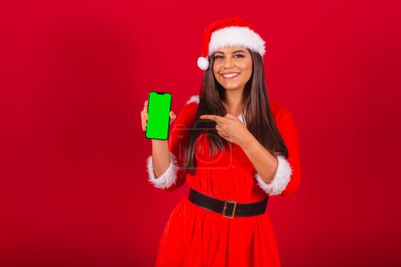 Téléchargez les photos : Belle Brésilienne vêtue de vêtements de Noël, le Père Noël. tenant smartphone montrant écran chromé vert. - en image libre de droit