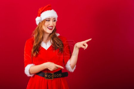 Photo for Beautiful Brazilian red-haired woman, dressed in Christmas clothes, Santa Claus. showing something to the right. - Royalty Free Image
