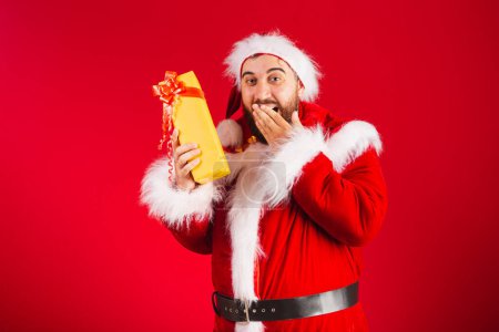 Foto de Hombre brasileño, vestido con ropa de Santa Claus, dudando de lo que hay en el regalo amarillo, duda, sorpresa. - Imagen libre de derechos