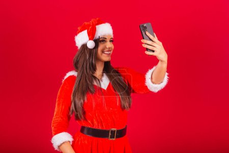 Photo for Beautiful Brazilian woman dressed in Christmas clothes, Santa Claus. holding smartphone taking self portrait. - Royalty Free Image