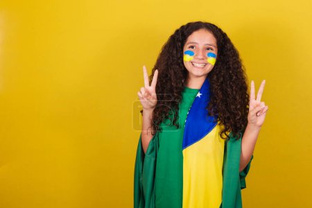 Téléchargez les photos : Brésilien, fille caucasienne, expression de paix et d'amour, doigts levés, pose heureuse pour la photo. Coupe du Monde. Jeux olympiques. fan de football - en image libre de droit