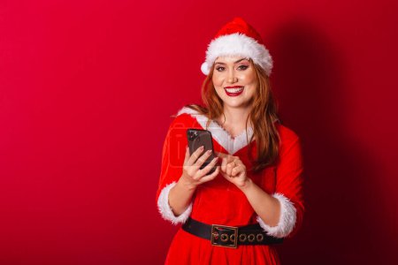 Photo for Beautiful Brazilian red-haired woman, dressed in Christmas clothes, Santa Claus. holding smartphone, typing. - Royalty Free Image