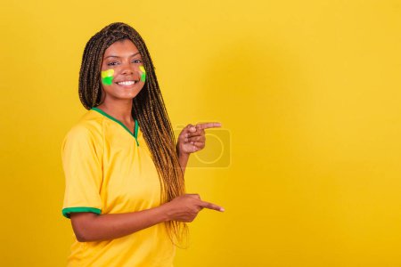 Foto de Mujer negra joven aficionada al fútbol brasileño. señalando con el dedo hacia la derecha, sonriendo, foto publicitaria. - Imagen libre de derechos