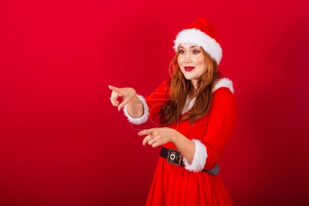Photo for Beautiful Brazilian red-haired woman, dressed in Christmas clothes, Santa Claus. pointing at something in the distance. - Royalty Free Image