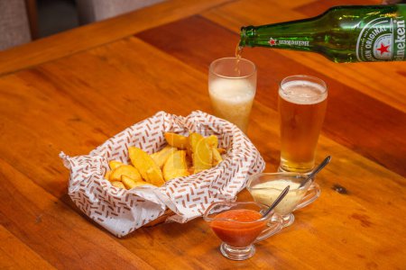 Téléchargez les photos : Des frites rustiques, une collation brésilienne typique, servies avec de la mayonnaise et de la sauce chili. Servi avec la célèbre bière Heineken - en image libre de droit