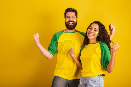 Téléchargez les photos : Couple de supporters brésiliens de football, vêtus aux couleurs de la nation, femme noire, homme caucasien. Torsion et vibration. - en image libre de droit