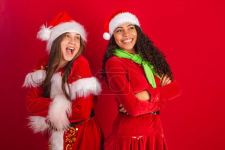 Foto de Dos amigas brasileñas, vestidas con ropa navideña, santa claus, con los brazos cruzados sonriendo con optimismo. - Imagen libre de derechos