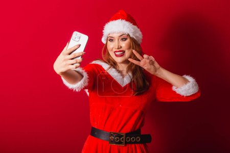 Photo for Beautiful Brazilian red-haired woman, dressed in Christmas clothes, Santa Claus. taking self portrait through smartphone. - Royalty Free Image
