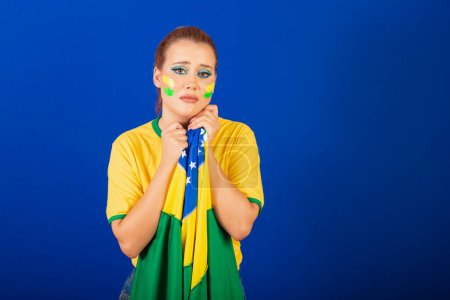 Photo for Caucasian woman, redhead, brazil soccer fan, brazilian, blue background, sad and upset with flag in hands - Royalty Free Image