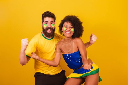 Foto de Pareja de aficionados brasileños al fútbol, vestidos con los colores de Brasil, mujer negra, hombre caucásico. gritando sí, celebrando la victoria - Imagen libre de derechos