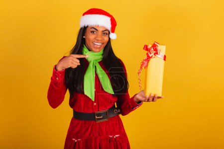 Téléchargez les photos : Belle Brésilienne noire vêtue de vêtements de Noël, Père Noël claus, pointant du doigt un cadeau jaune, doute, - en image libre de droit