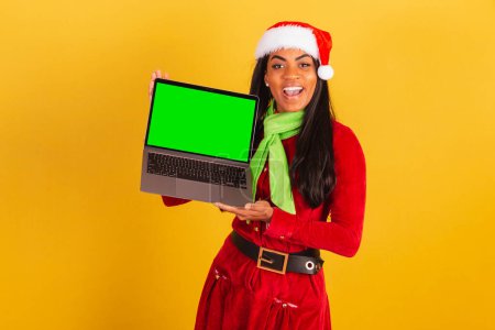 Téléchargez les photos : Belle femme brésilienne noire, vêtue de vêtements de Noël, Père Noël claus, carnet et montrant écran chromé vert. - en image libre de droit