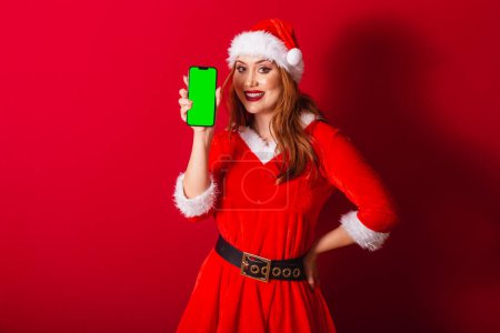 Téléchargez les photos : Belle femme rousse brésilienne, vêtue de vêtements de Noël, le Père Noël. montrant smartphone avec écran chromatique vert. - en image libre de droit