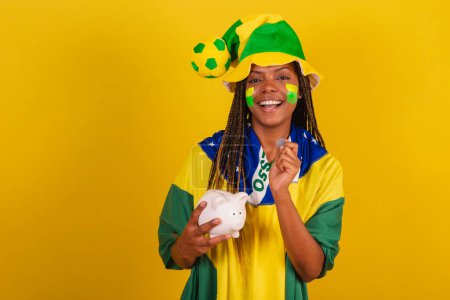 Téléchargez les photos : Femme noire jeune fan de football brésilien. tenant tirelire et monnaie. concept de finance, économie. - en image libre de droit