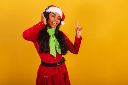 Téléchargez les photos : Belle femme brésilienne noire, vêtue de vêtements de Noël, Père Noël claus, écoutant de la musique dans les écouteurs. - en image libre de droit