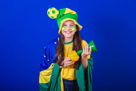 Téléchargez les photos : Jeune fille, fan de football du Brésil. habillé en chapeau et drapeau. faire du bruit avec corne - en image libre de droit